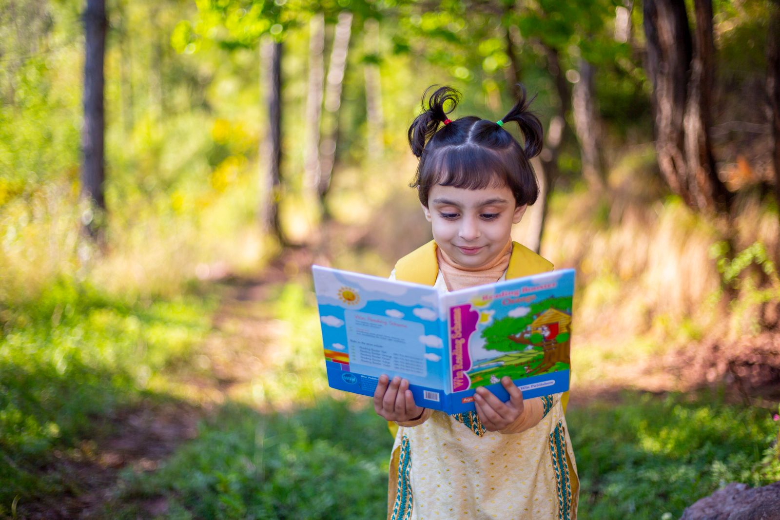 A young girl standing among trees, smiling as she reads a picture book